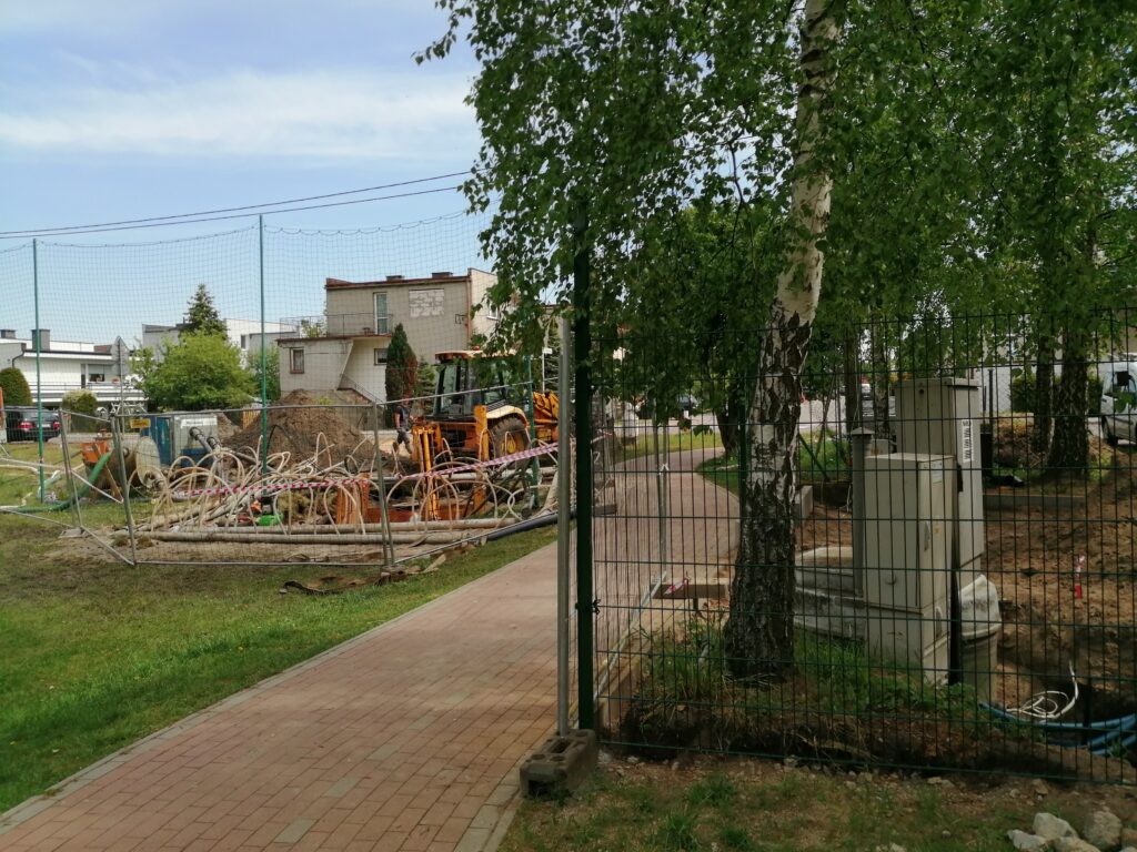 Plac budowy przepompowni ścieków „Wiśniowa” za ogrodzeniem, w tle zabudowa.